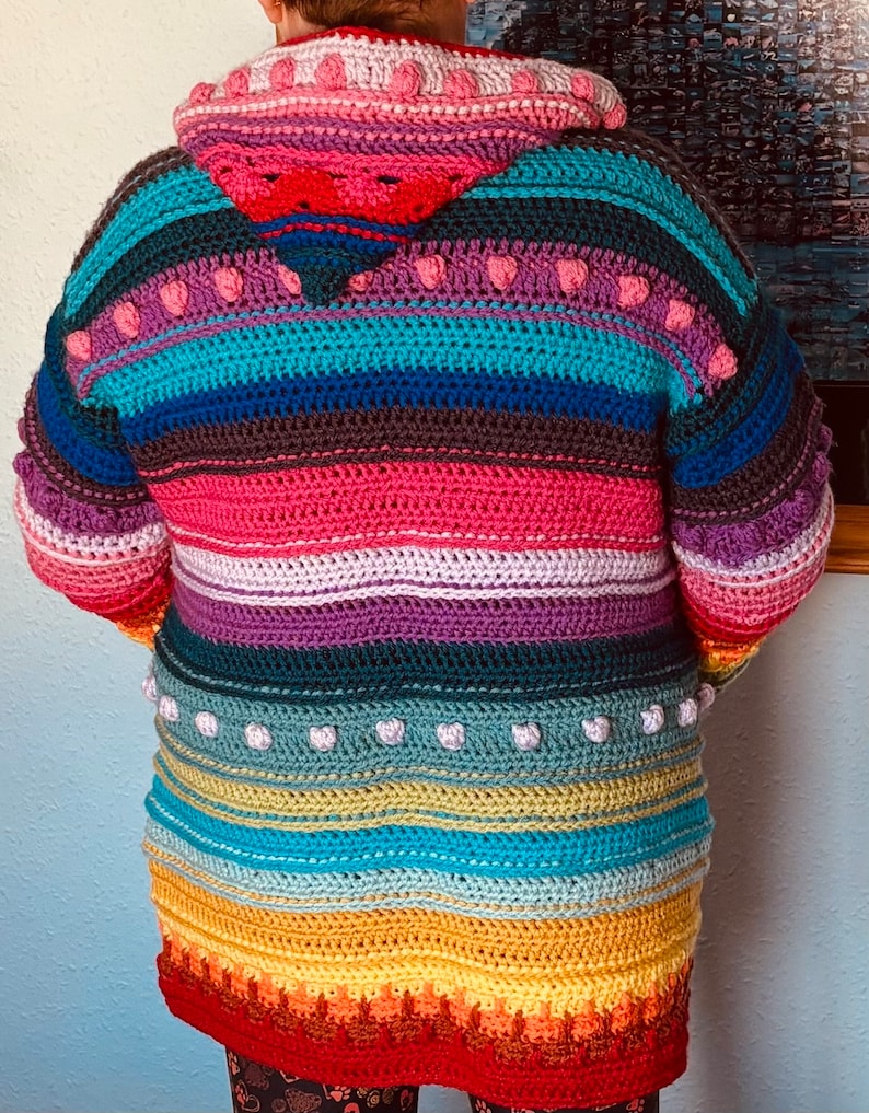 Puede incluir: Un c&aacute;rdigan de crochet a rayas de colores con capucha. El c&aacute;rdigan presenta un esquema de color arco&iacute;ris con rayas rosas, moradas, azules, verdes, amarillas, naranjas y rojas. La capucha est&aacute; forrada de rosa y tiene un ribete de crochet blanco.