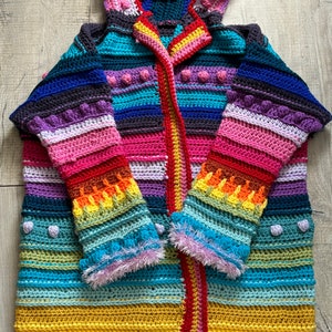 Puede incluir: Un c&aacute;rdigan de crochet a rayas de colores con capucha. El c&aacute;rdigan tiene una combinaci&oacute;n de colores rojo, naranja, amarillo, verde, azul, morado y rosa. El c&aacute;rdigan tiene una cremallera y est&aacute; hecho con una variedad de puntos de crochet.