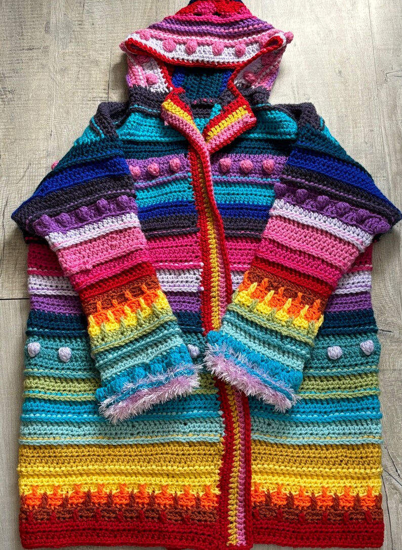 Puede incluir: Un c&aacute;rdigan de crochet a rayas de colores con capucha. El c&aacute;rdigan tiene una combinaci&oacute;n de colores rojo, naranja, amarillo, verde, azul, morado y rosa. El c&aacute;rdigan tiene una cremallera y est&aacute; hecho con una variedad de puntos de crochet.