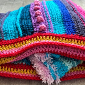 Puede incluir: Una manta de crochet colorida con rayas de color rosa, azul, turquesa, morado y rojo. La manta tiene un borde texturizado con flecos.