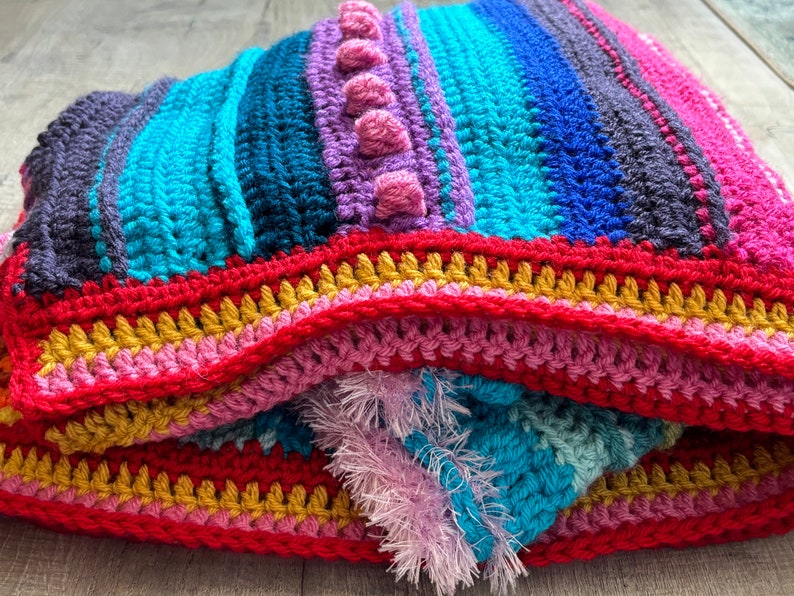 Puede incluir: Una manta de crochet colorida con rayas de color rosa, azul, turquesa, morado y rojo. La manta tiene un borde texturizado con flecos.