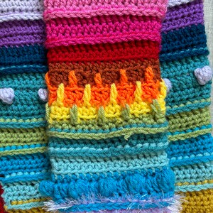 Puede incluir: Una manta de crochet colorida con rayas de color morado, rosa, rojo, naranja, amarillo, verde, azul y marr&oacute;n. La manta tiene un borde de flecos.
