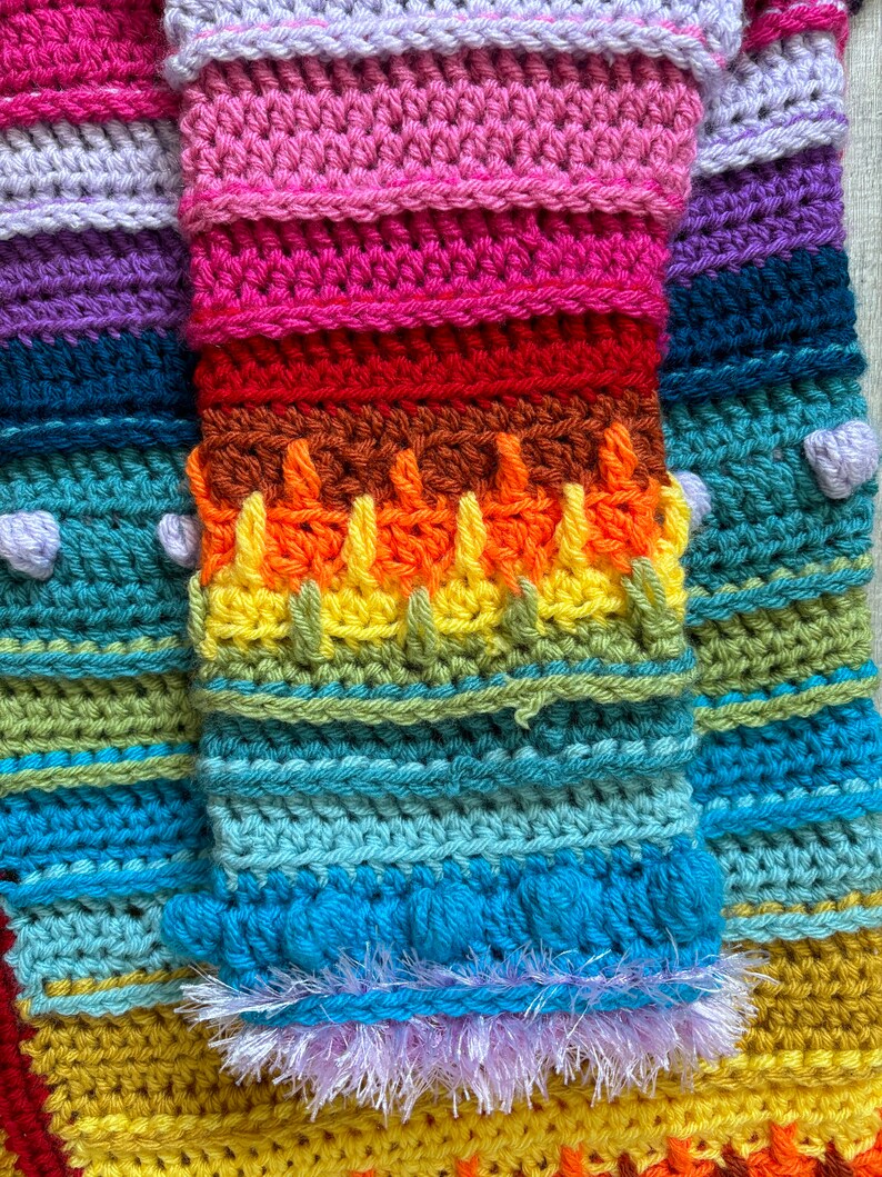 Puede incluir: Una manta de crochet colorida con rayas de color morado, rosa, rojo, naranja, amarillo, verde, azul y marr&oacute;n. La manta tiene un borde de flecos.