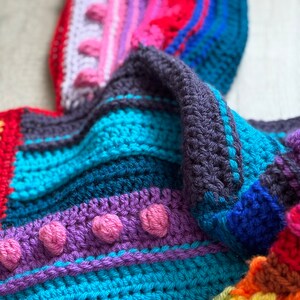 Puede incluir: Una manta de crochet colorida con rayas de rojo, rosa, morado, azul y amarillo. La manta tiene una superficie texturizada con peque&ntilde;os bultos y est&aacute; doblada de una manera que muestra los diferentes colores.