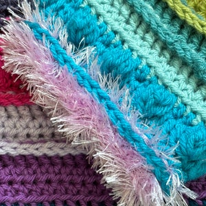 Puede incluir: Primer plano de una manta de ganchillo multicolor con un ribete rosa peludo. La manta est&aacute; hecha con diferentes tonos de azul, verde, morado y gris.