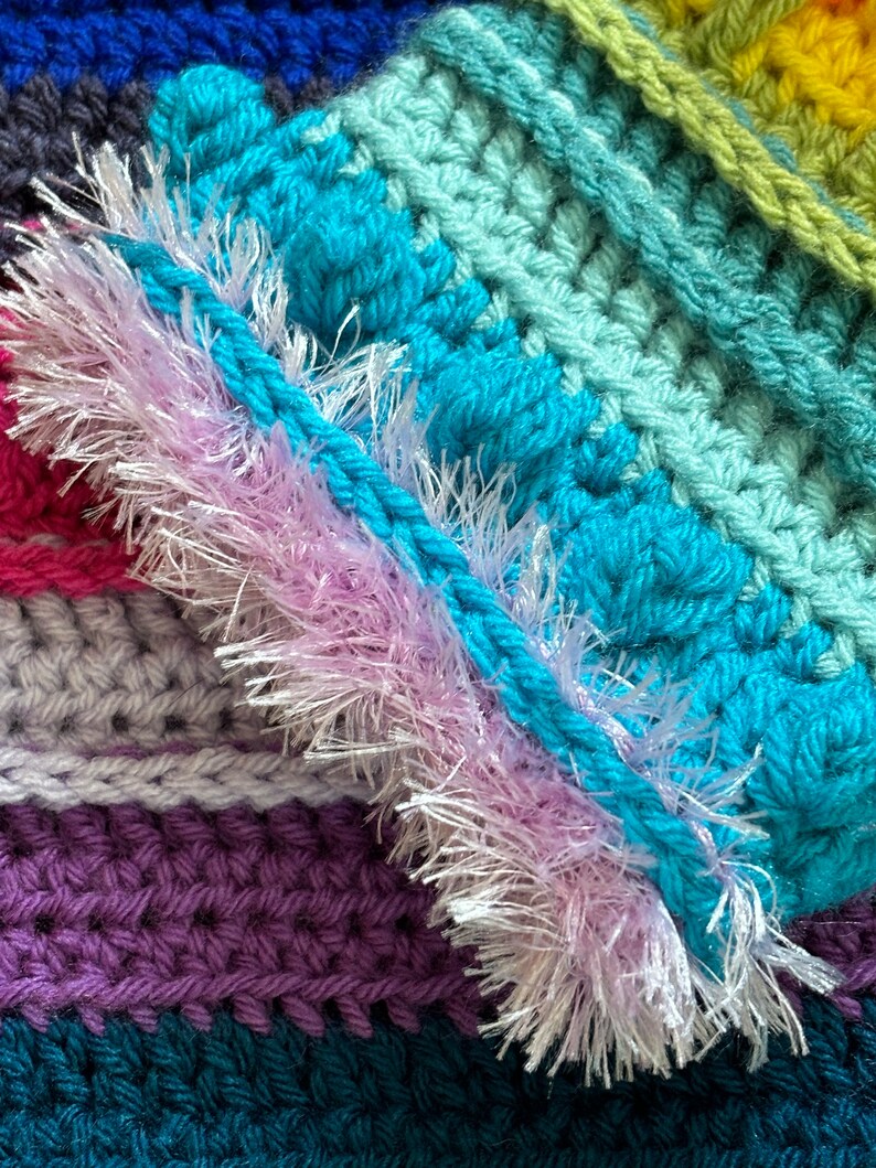 Puede incluir: Primer plano de una manta de ganchillo multicolor con un ribete rosa peludo. La manta est&aacute; hecha con diferentes tonos de azul, verde, morado y gris.