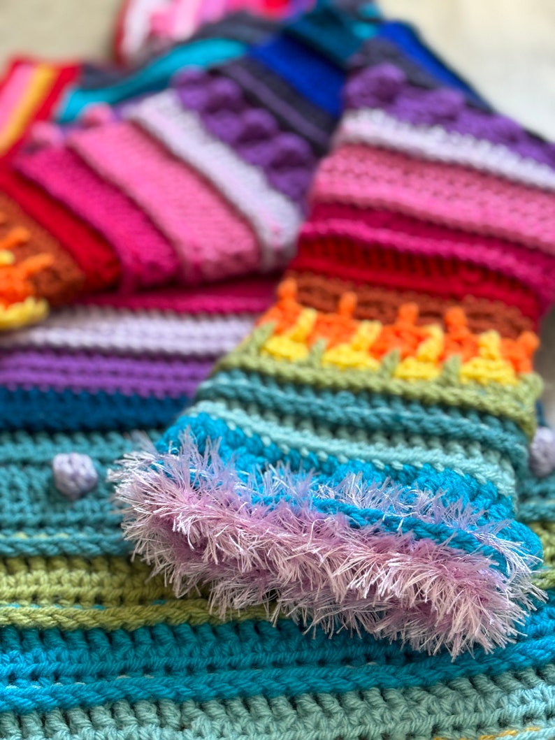 Puede incluir: Una bufanda de crochet colorida con un patr&oacute;n de arco&iacute;ris y un borde esponjoso azul y morado.