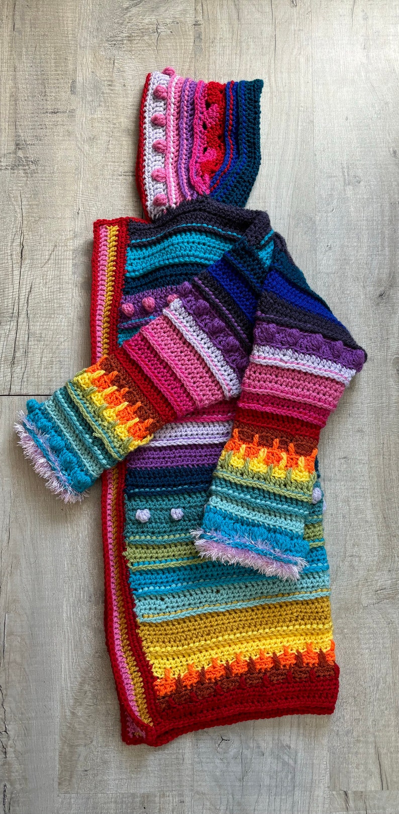 Puede incluir: Un c&aacute;rdigan de ganchillo colorido con capucha, con rayas de color rosa, morado, azul, verde, amarillo y rojo. El c&aacute;rdigan tiene un dise&ntilde;o &uacute;nico con puntadas texturizadas y pompones.