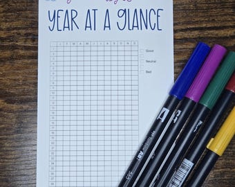 May include: A white Year at a Glance journal with a grid layout and the text "Journals By Luck" and "YEAR AT A GLANCE" in purple and blue. Five Tombow ABT markers in blue, purple, green, red, and yellow are next to the journal.