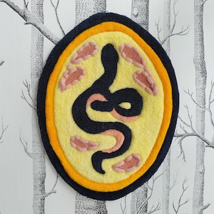 Pode incluir: Um remendo circular de feltro amarelo e laranja com um desenho de cobra preta. A cobra é contornada com feltro rosa e tem um desenho de feltro rosa no interior.