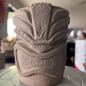 K&ouml;nnte beinhalten: Unglasierter Keramik-Tiki-Becher in Hellbraun. Der Becher hat ein detailliertes, geschnitztes Design eines Tiki-Gesichts mit breitem Mund, mandelf&ouml;rmigen Augen und strukturierter Oberfl&auml;che. Der Becher ist zylindrisch mit flachem Boden.