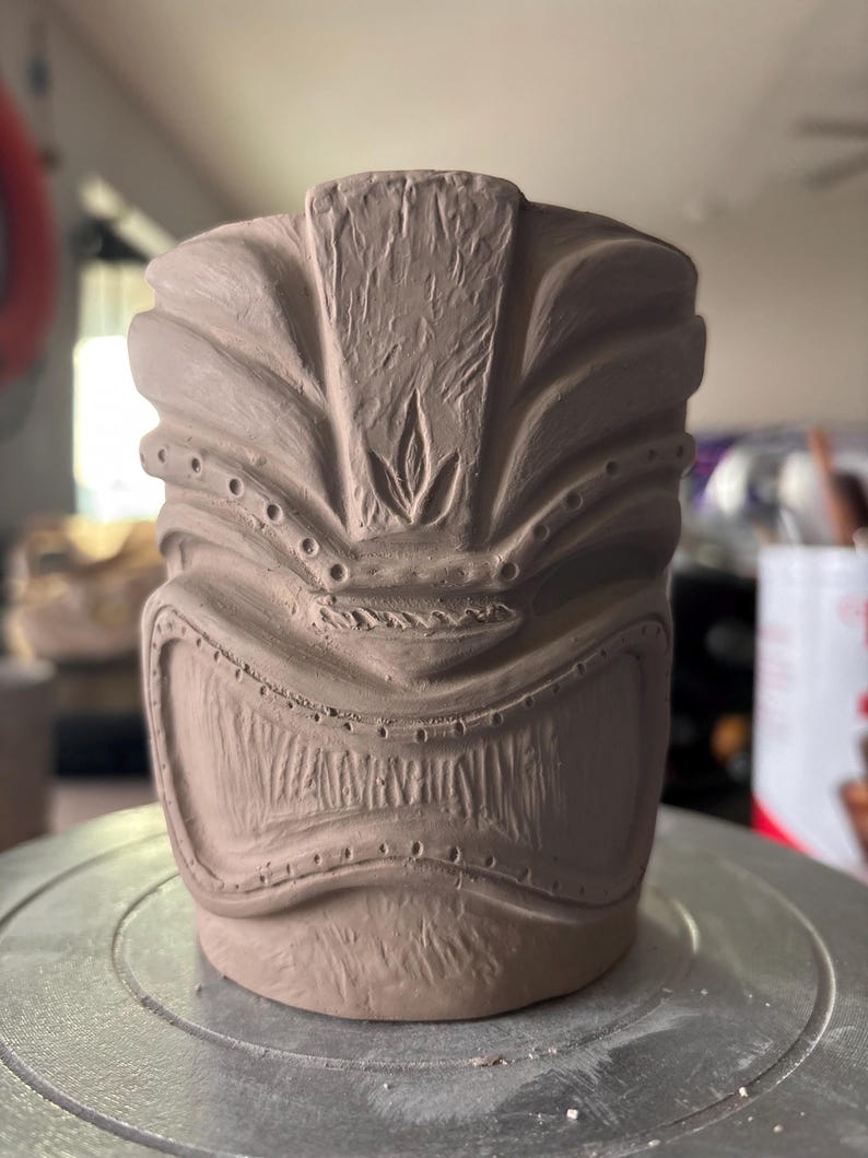 K&ouml;nnte beinhalten: Unglasierter Keramik-Tiki-Becher in Hellbraun. Der Becher hat ein detailliertes, geschnitztes Design eines Tiki-Gesichts mit breitem Mund, mandelf&ouml;rmigen Augen und strukturierter Oberfl&auml;che. Der Becher ist zylindrisch mit flachem Boden.