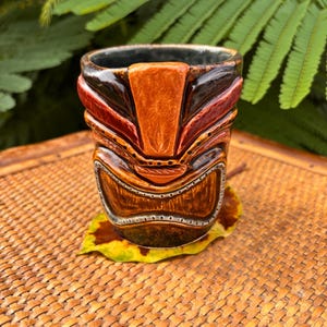 K&ouml;nnte beinhalten: Ein handgefertigter Tiki-Becher mit detailliertem Gesichtsdesign. Der Becher hat braune, rote und orangefarbene T&ouml;ne mit dunklem Innenraum. Er liegt auf einem gelb-braunen Blatt, vor einem gewebten Hintergrund und gr&uuml;nem Laub.