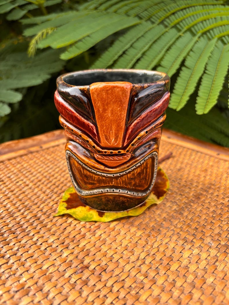 K&ouml;nnte beinhalten: Ein handgefertigter Tiki-Becher mit detailliertem Gesichtsdesign. Der Becher hat braune, rote und orangefarbene T&ouml;ne mit dunklem Innenraum. Er liegt auf einem gelb-braunen Blatt, vor einem gewebten Hintergrund und gr&uuml;nem Laub.
