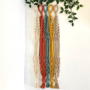May include: Five macrame plant hangers in different colours: cream, orange, teal, yellow, and beige. The hangers are made with cotton cord and have a knotted design.