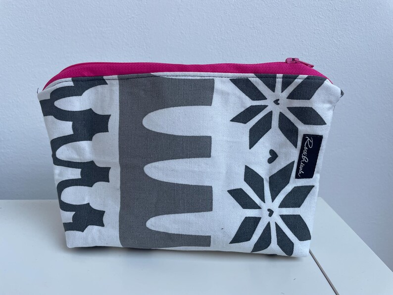 Puede incluir: Una bolsa blanca con cremallera con un patr&oacute;n geom&eacute;trico blanco y negro. La bolsa tiene una cremallera rosa y una etiqueta negra con el texto "Rosa & Black" en ella.