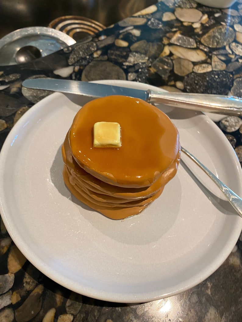 May include: A stack of four golden brown pancakes with a pat of butter on top. The pancakes are on a white plate with a silver knife and fork next to them.