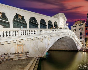 May include: A white stone bridge arches over a canal in Venice, Italy. The bridge has a walkway with a white stone railing. The sky is a vibrant pink and purple.