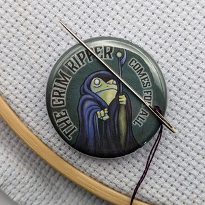 May include: A round button with a green background and a frog wearing a dark blue robe and holding a staff. The text on the button reads "This Grim Ripper Comes For All".
