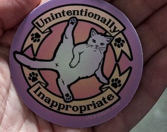 Puede incluir: Una pegatina redonda con fondo morado y un gato blanco con la pata levantada en el aire. El gato está rodeado de una pancarta amarilla con el texto "Unintentionally Inappropriate" y huellas de patas. La pegatina tiene el texto "LOLA EDGAR.COM" en la parte inferior.