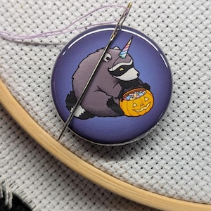 Op de afbeelding: Een ronde naaldmagneet met een paarse achtergrond en een illustratie van een grijze wasbeer met een eenhoornhoorn die een oranje pompoen vol gekleurde snoepjes vasthoudt, inclusief een zilverkleurige metalen naald.