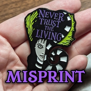 May include: Black and white enamel pin with a green and purple design. The pin features a character with black hair and a white face. The text on the pin reads "Never Trust The Living". The word "Misprint" is written in purple letters below the pin.