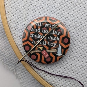 Könnte beinhalten: Nahaufnahme eines runden Knopfes mit einem rot-schwarzen Muster. Der Knopf trägt den Text "All work and no play makes Jack a dull boy." und ein kleines rotes Etikett mit dem Text "Room No. 237".