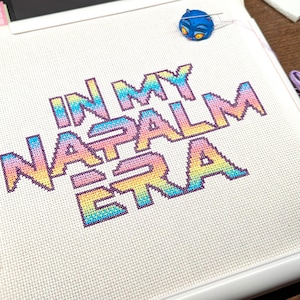 May include: Cross-stitch project in a white frame, featuring the text "IN MY NAPALM ERA" in rainbow colors. Includes embroidery floss, scissors, and a blue pin. A creative craft project.