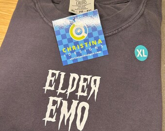 May include: A dark gray t-shirt with white text that reads "ELPER EMO". The shirt is size XL and has a tag that says "Christina Designs" and a website address.