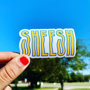 May include: A white sticker with the word "SHEESH" written in yellow and blue letters. The sticker is being held by a hand with red nail polish.