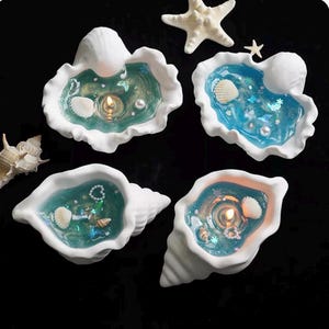 May include: Four white ceramic shell-shaped containers filled with blue and green gel wax. Each contains a lit wick, small embedded seashells, pearls, and glitter. Two starfish are placed separately among the shell containers.