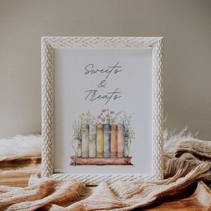 Puede incluir: Impresión enmarcada en blanco con el texto "Sweets & Treats" en escritura cursiva negra. La impresión presenta una ilustración de acuarela de una pila de libros coloridos con flores y vegetación.