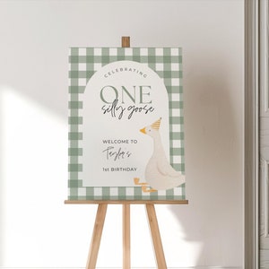 May include: A wooden easel displays a sign with a green and white checkered border. The sign reads "Celebrating One Silly Goose" and "Welcome to Taylor's 1st Birthday" with a cartoon goose illustration wearing a party hat.