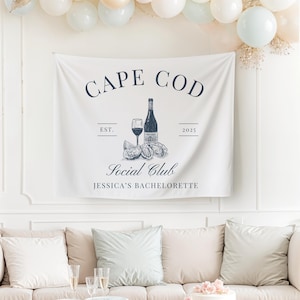 May include: A white banner with blue text that reads "Cape Cod Social Club Jessica's Bachelorette Est. 2025". The banner features a drawing of a bottle of wine, a glass of wine, and two oysters.