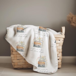 May include: A white blanket with a repeating pattern of stacked books and floral arrangements. The text "JUST A GIRL who loves books" is printed on the blanket. The blanket is draped over a woven basket.