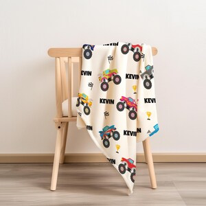 May include: A white and cream-colored blanket with a monster truck pattern. The blanket features a variety of monster trucks in different colors, including yellow, red, blue, and green. The blanket also has the name "Kevin" printed on it multiple times.