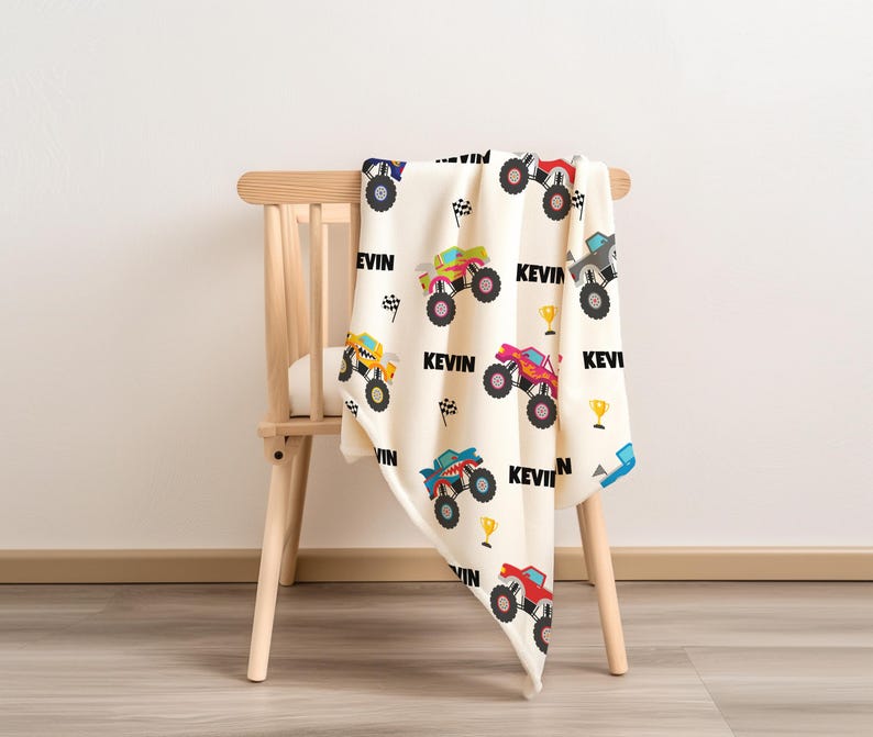 May include: A white and cream-colored blanket with a monster truck pattern. The blanket features a variety of monster trucks in different colors, including yellow, red, blue, and green. The blanket also has the name "Kevin" printed on it multiple times.