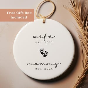 May include: A white ceramic ornament with a jute string for hanging. The ornament features the words "wife est. 2021" above a pair of baby footprints and the words "mommy est. 2025". A gift box is included.