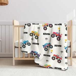 May include: White baby blanket with a repeating pattern of monster trucks in various colors, including blue, yellow, red, and pink. The blanket features the name "Kevin" repeated throughout the design.