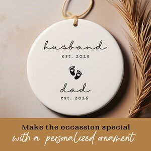 May include: A white ceramic ornament with the words "husband est. 2023" and "dad est. 2026" in black script. The ornament features a pair of black footprints between the words. A beige string is attached for hanging.
