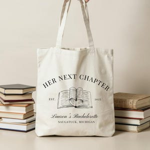 May include: A white canvas tote bag with black text that reads "Her Next Chapter" with an illustration of an open book. The text also includes "Est. 2025" and "Lauren's Bachelorette, Saugatuck, Michigan".
