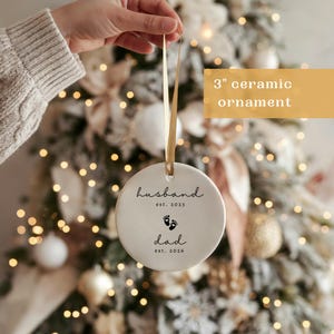 May include: A 3-inch ceramic ornament with a gold ribbon. The white ornament has the words "husband est. 2023" and "dad est. 2026" with baby footprints. The background is a decorated Christmas tree.