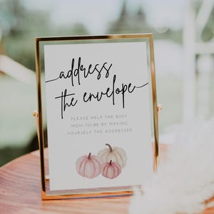 May include: A gold-framed sign with the text "address the envelope" and a watercolor illustration of three pink and white pumpkins. The sign reads "Please help the busy mom-to-be by making yourself the addressee."