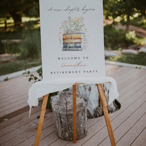 May include: A white sign with the text "A new chapter begins" and a watercolour illustration of a stack of books with flowers on top. The sign also says "Welcome to Samantha's Retirement Party". The sign is on a wooden easel.