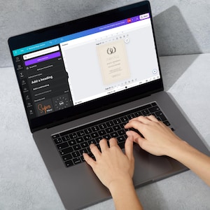 May include: A person is using a laptop computer to design a wedding invitation. The invitation is on the screen and features a laurel wreath design. The laptop has a gray keyboard and a black trackpad.