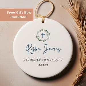 May include: A white ceramic ornament with a cross design, a wreath, and the name "Ryker James" in blue script. The text "Dedicated to our Lord" and a date are also included. A beige string is attached for hanging.