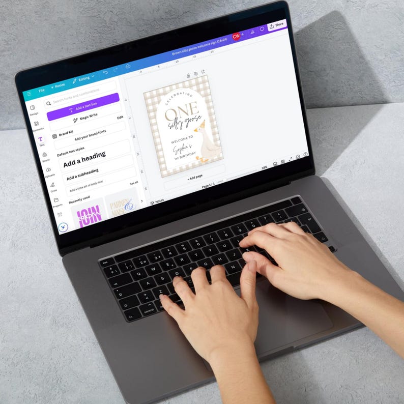 May include: A laptop computer with a white screen displaying a design for a birthday party invitation. The design features a white goose with a gold crown and the text "Celebrating One Silly Goose". The text "Welcome to Sophia's 1st Birthday" is also included in the design.