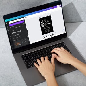 May include: A person is using a laptop computer to design a black and white birthday invitation with the text "Bad Two The Bone" and a skeleton hand making a peace sign. The invitation is for a 2nd birthday.