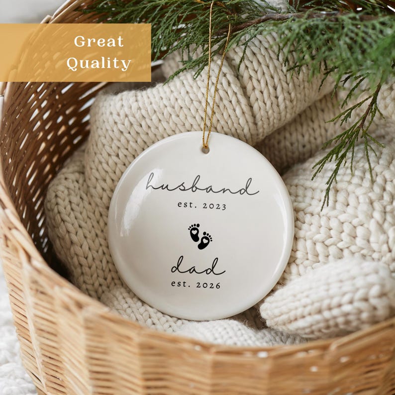 May include: A white ceramic Christmas ornament with the words "husband est. 2023" and "dad est. 2026" in black script. The ornament features two small black footprints and hangs from a gold string. The ornament is in a woven basket.