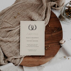May include: A wedding invitation with a rustic theme. The invitation features two horseshoes intertwined and the text "We invite you to celebrate our marriage" in cursive. The names Bailey Thompson and Stetson Rogers are printed in a bold font. The date and time of the wedding are listed as Saturday, August Thirty-first, Two Thousand Twenty-Five at Half Past Four in the Afternoon. The location is listed as 123 Anywhere St, Any City. The invitation concludes with "Reception to Follow."